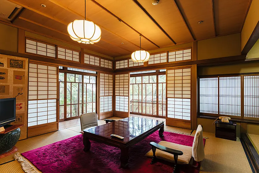 Daimaru Besso 大丸別荘 — Suita-no-yu communal bath interior with stone-lined pool, wide windows opening to garden trees, Futsukaichi Onsen. | Hotel Manga