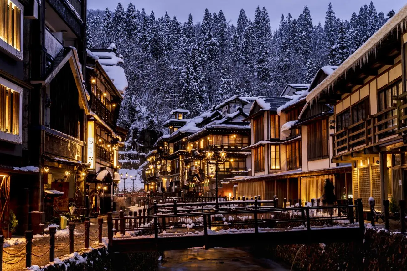 Ginzan Onsen gas-lit street at night in winter — snow on wooden ryokan rooftops, Ginzan River flowing between Taisho-era buildings Yamagata